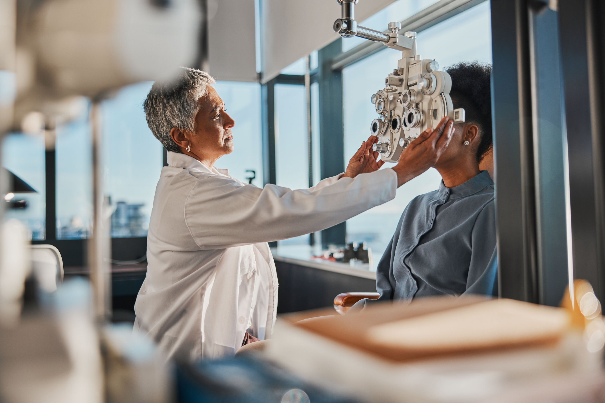 Woman sitting in an exam chair at the optometrist office, female optometrist holding phoropter up to her face for an exam